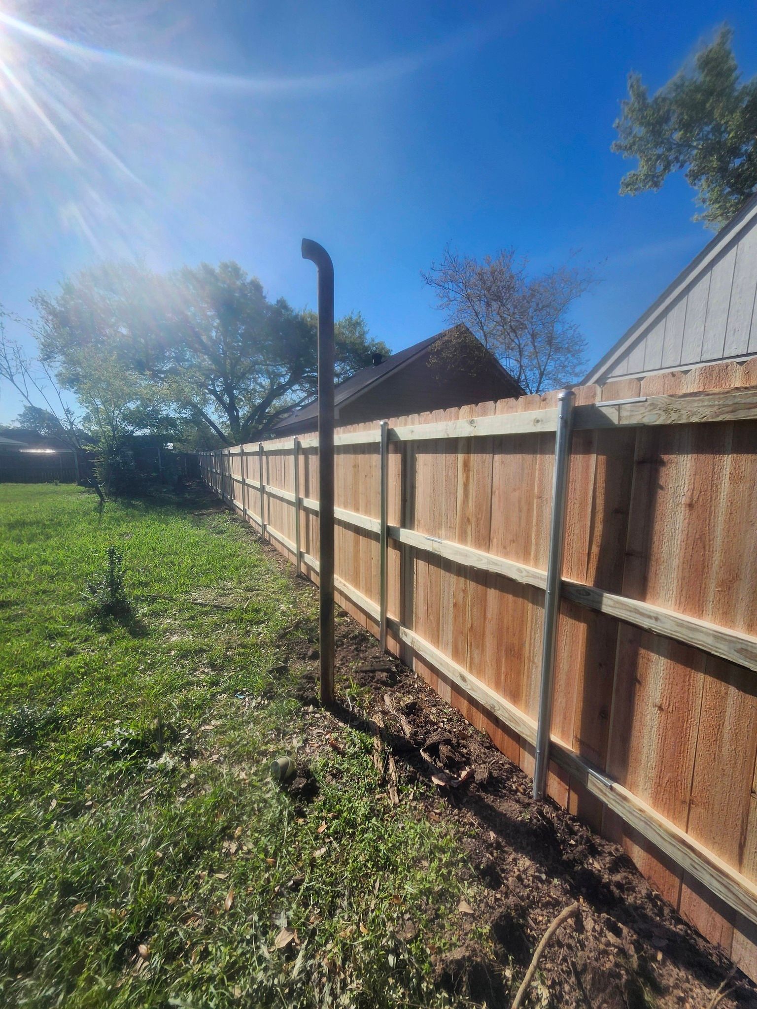 Wooden fence along a grassy yard under bright sun, with trees and a house roof in the background