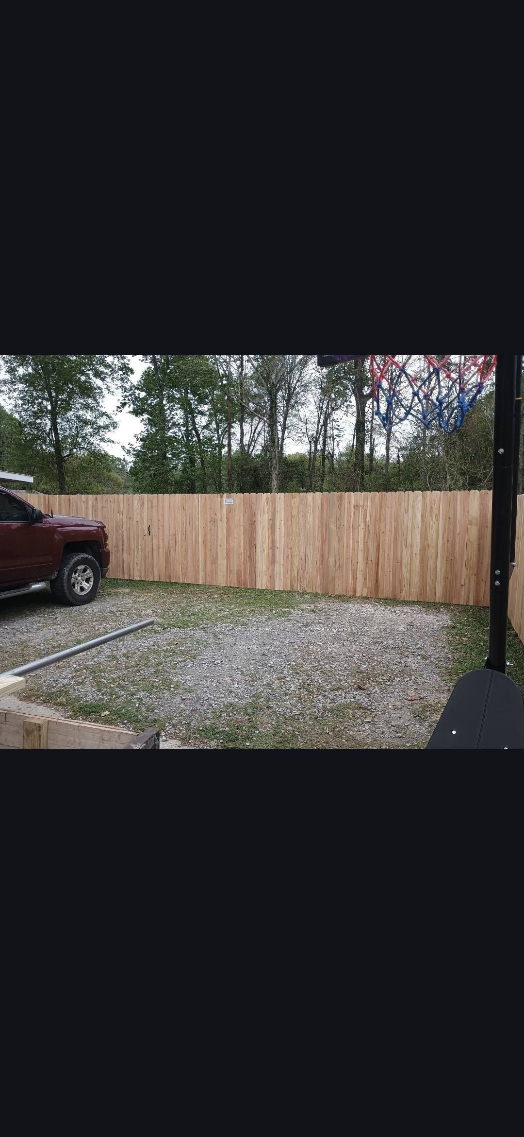 Wooden privacy fence beside a gravel driveway, with a red vehicle at left and trees beyond.