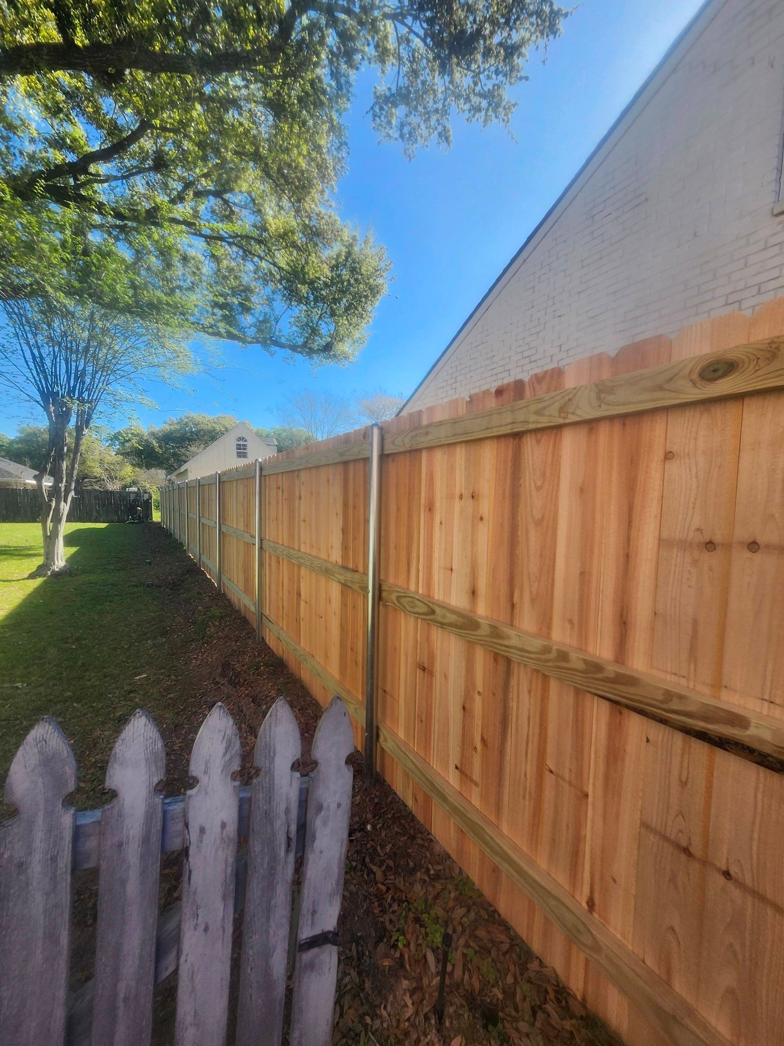 Wooden fence along a grassy side yard under a blue sky, with a weathered white picket fence in the foreground