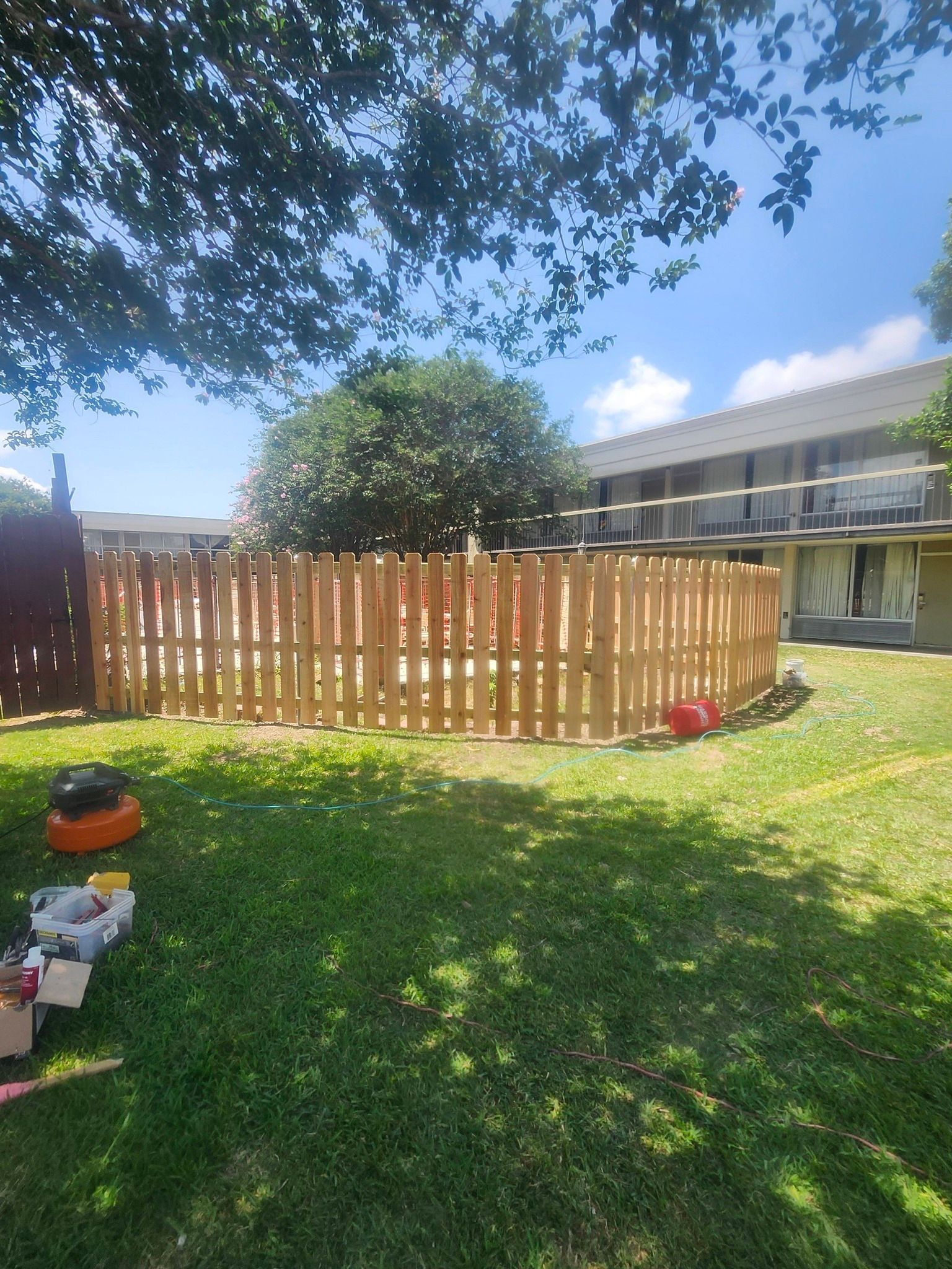 Backyard with wooden fence, green lawn, tree shade, and a house in the background.