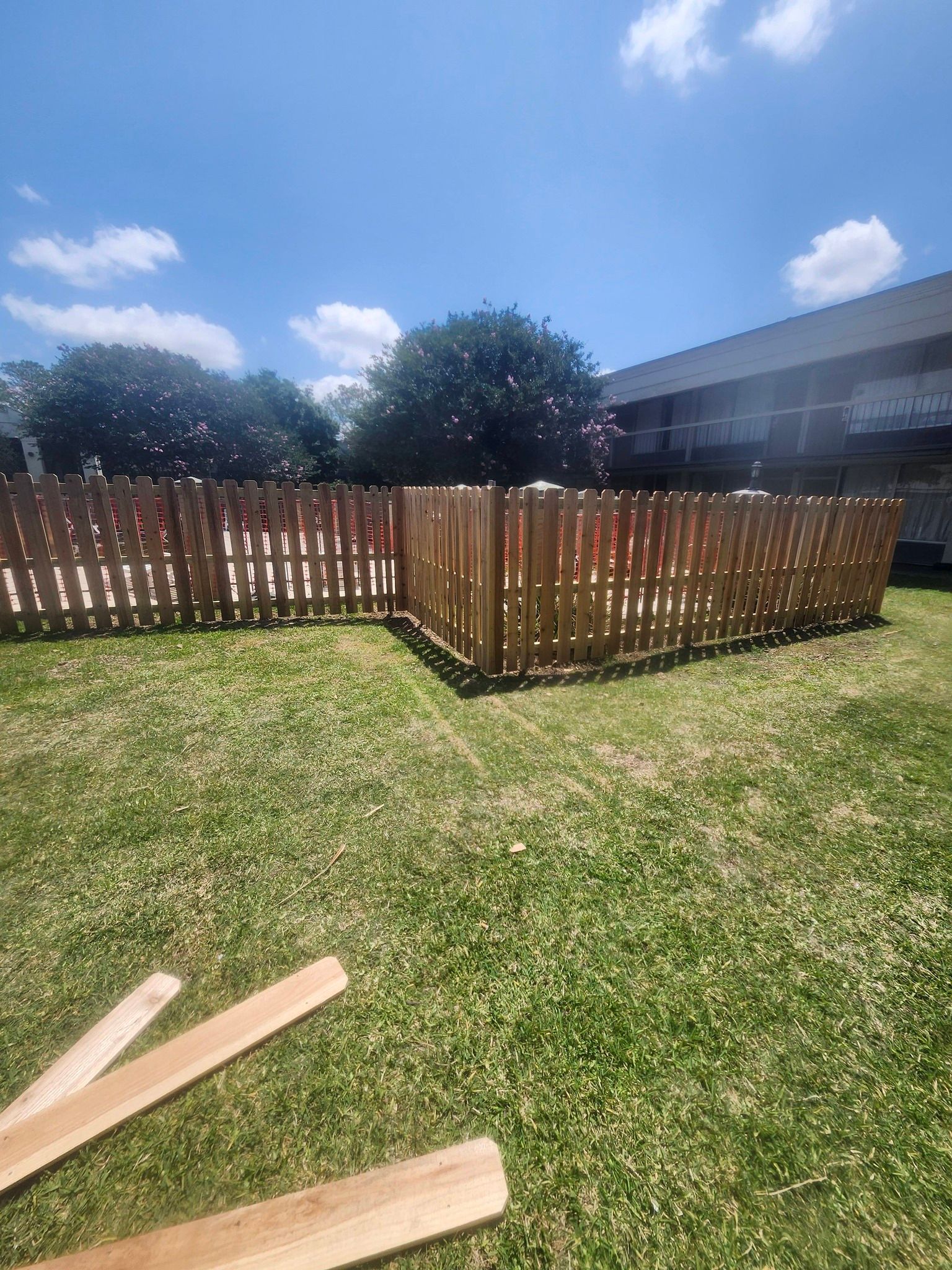 Backyard with wooden fence around a grass lawn under a blue sky, with two planks in the foreground.