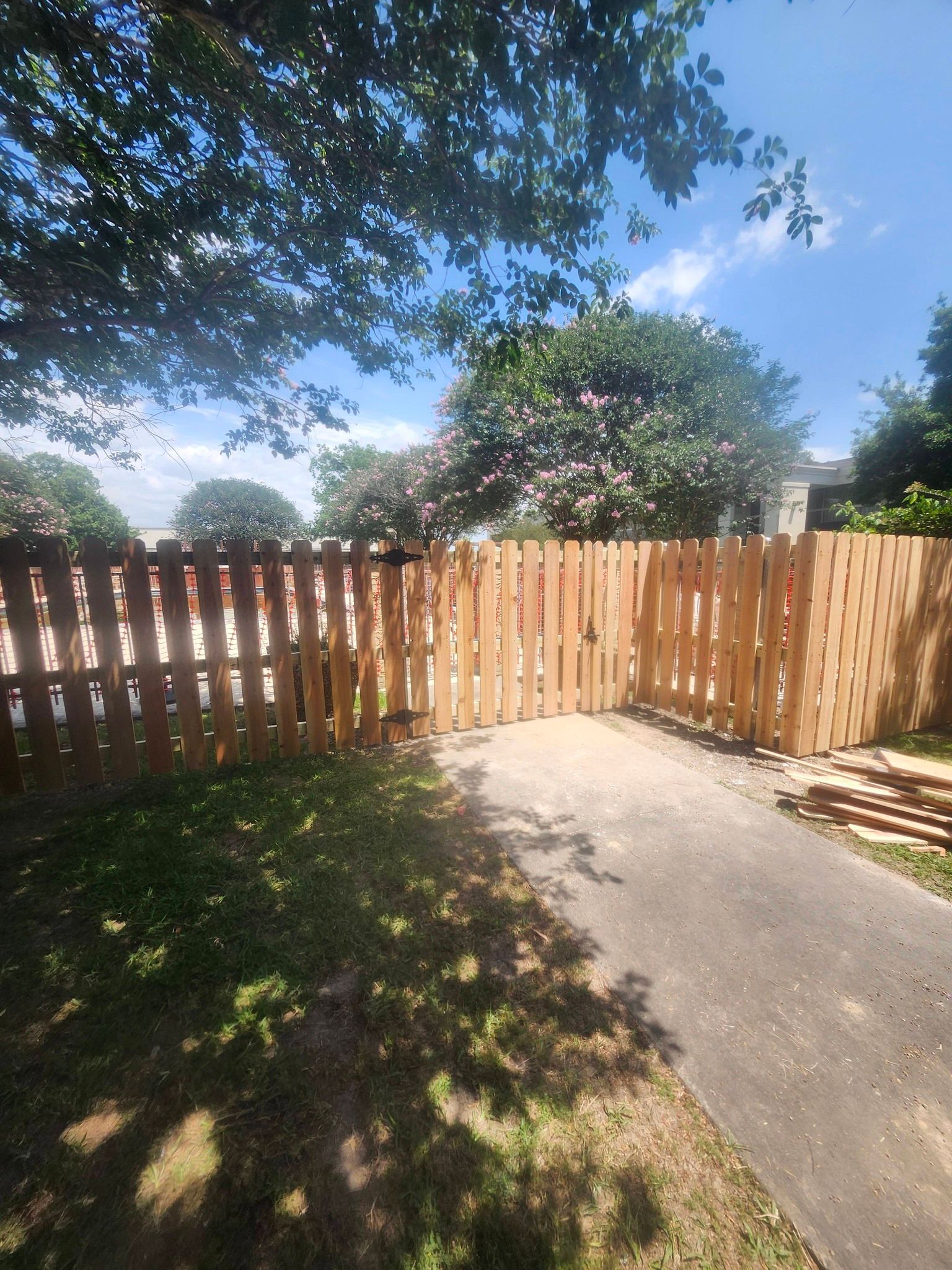 Backyard with wooden fence, concrete patio, and trees casting dappled shade under a bright blue sky