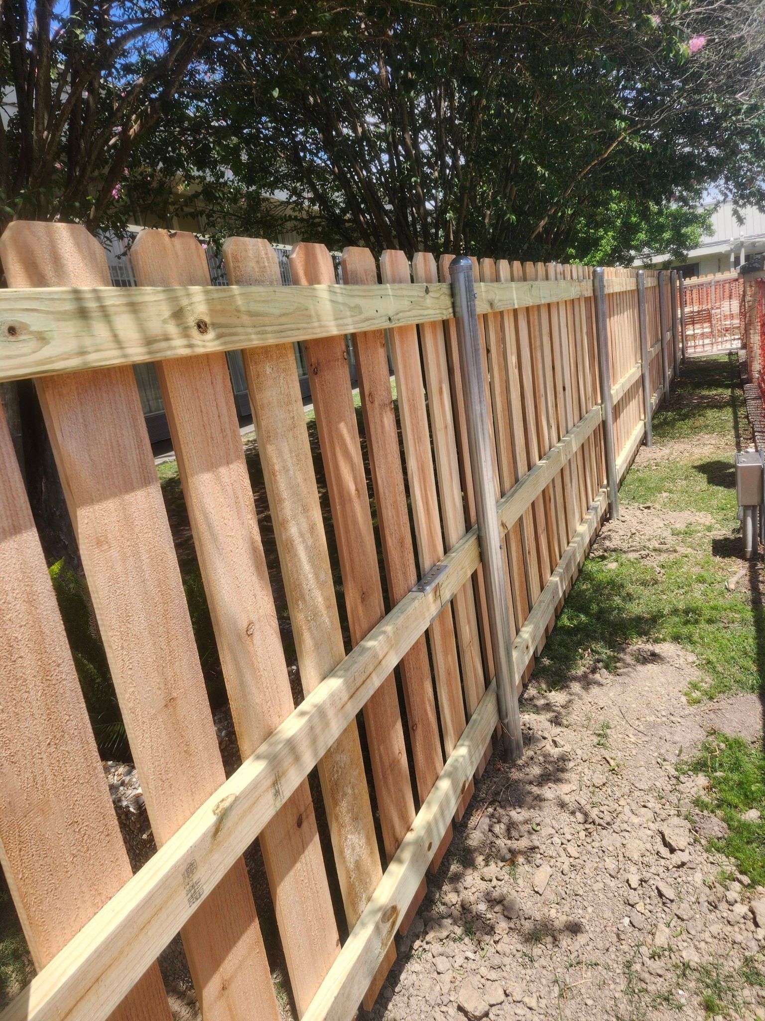 Wooden fence along a narrow dirt path, with trees overhead and a yard in the background