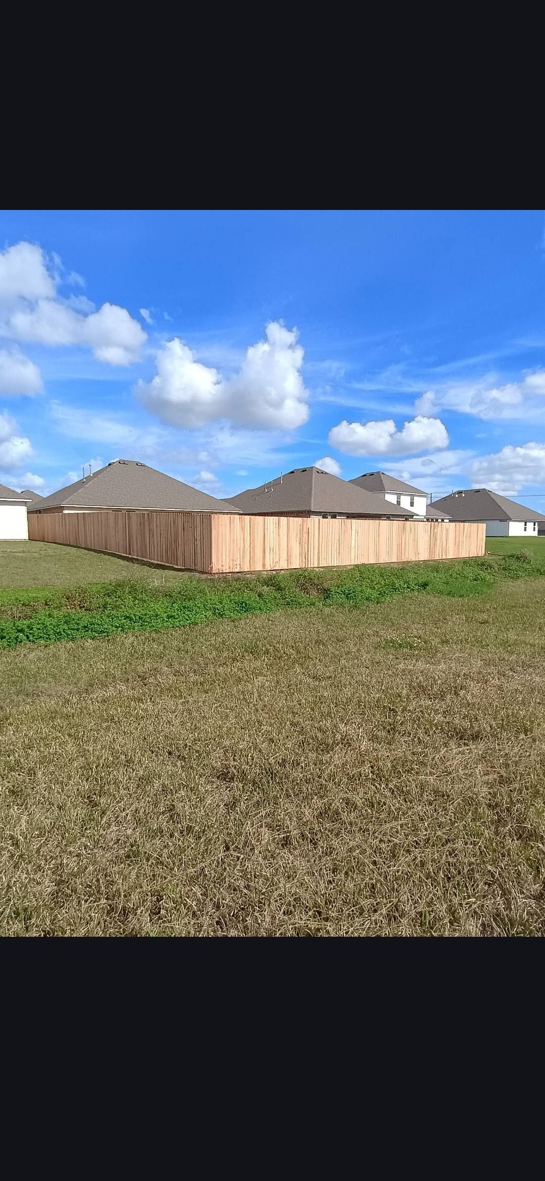 Row of tan houses with brown roofs beside a grassy field under a blue sky with clouds