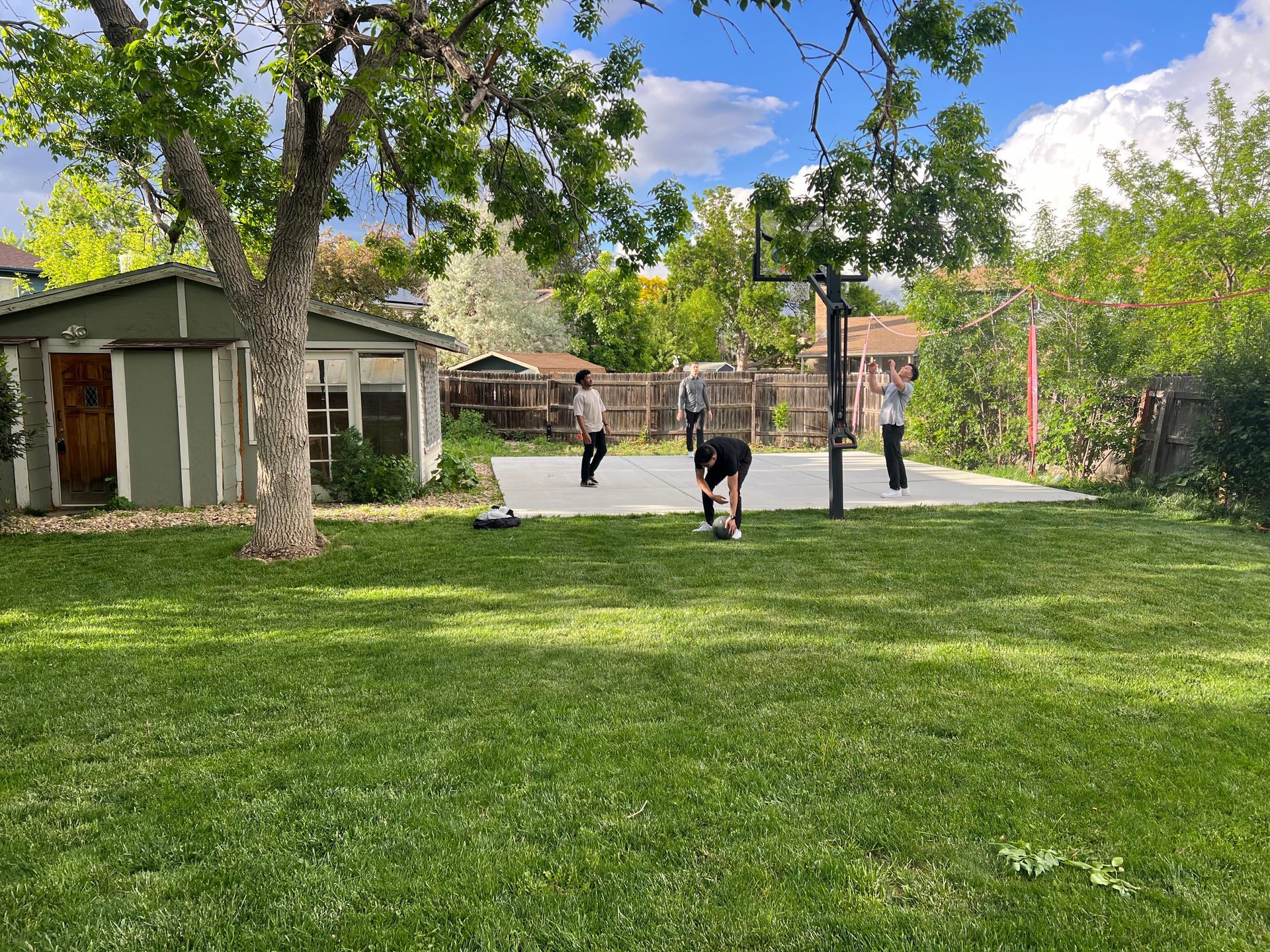 Sunny backyard with green lawn, trees, and two people near a patio and basketball hoop