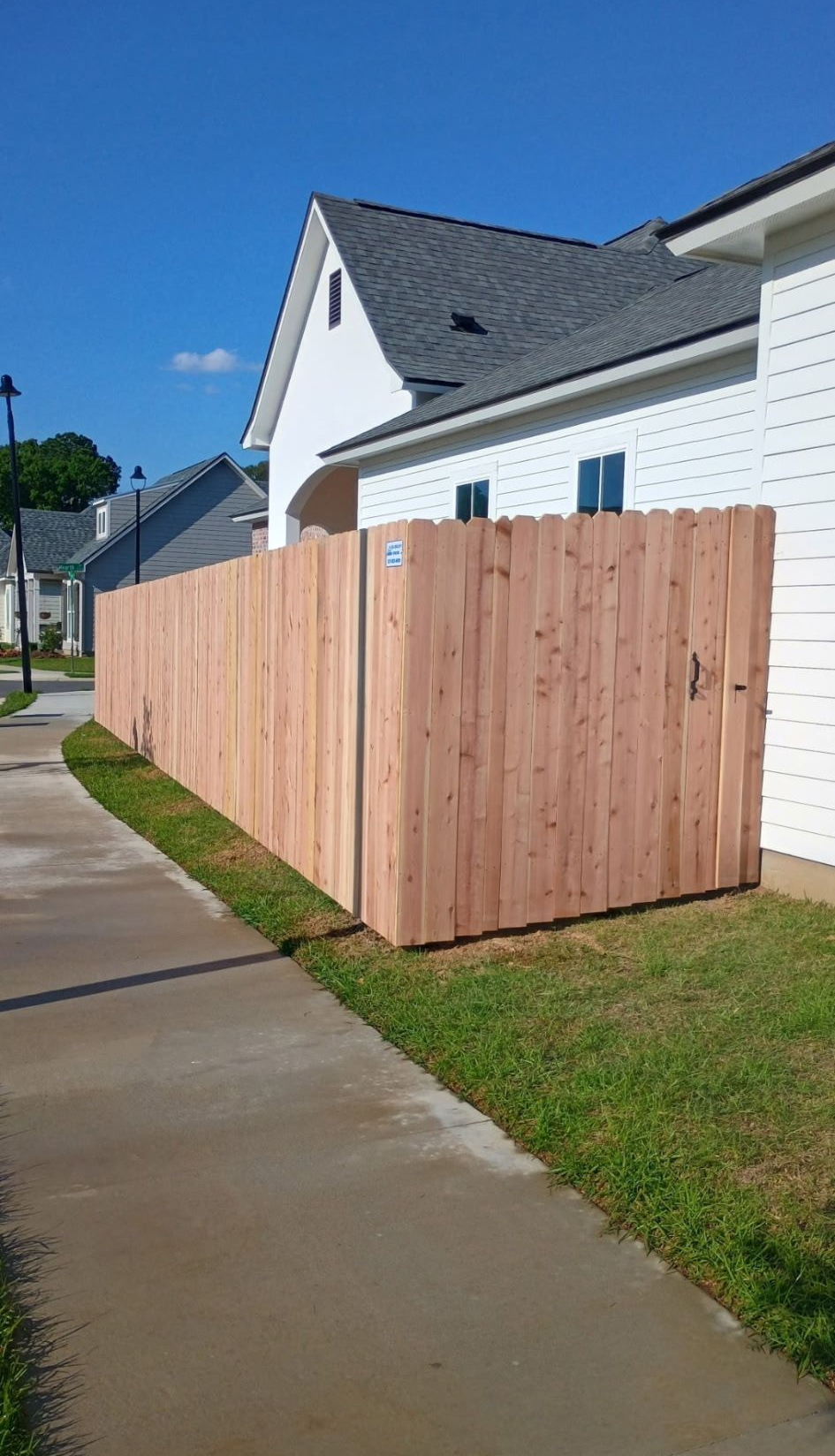 Wooden privacy fence beside a white house along a wet sidewalk and grassy verge.