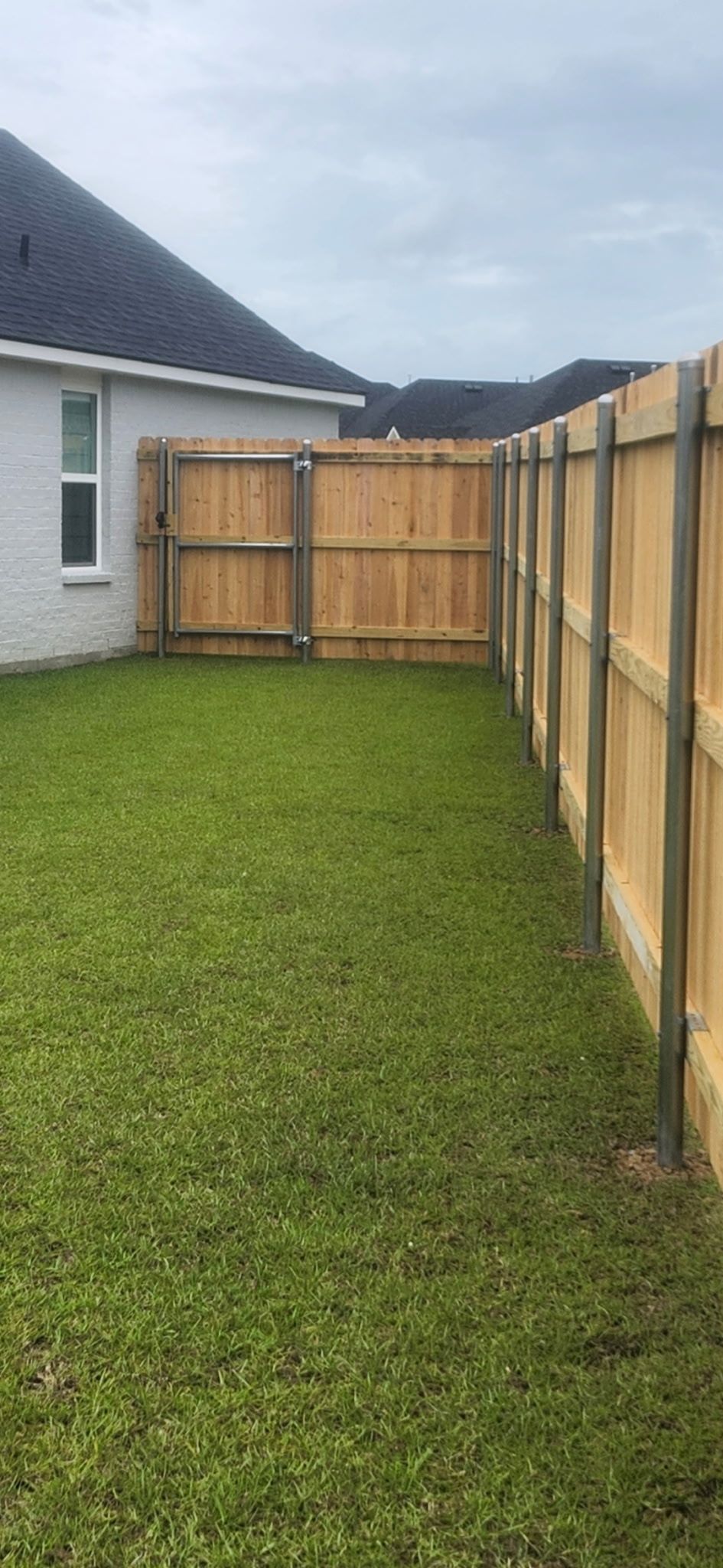 Narrow grassy side yard between a white house and wooden fence, with a gate at the end.