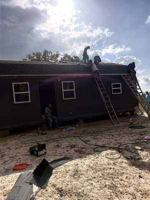 Workers on a roof repairing storm damage to a dark house, with a ladder leaning against it.