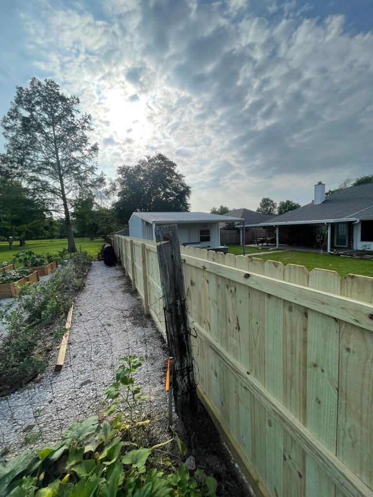 Gravel path beside a weathered wooden fence, with trees, lawn, and a cloudy sunlit sky.