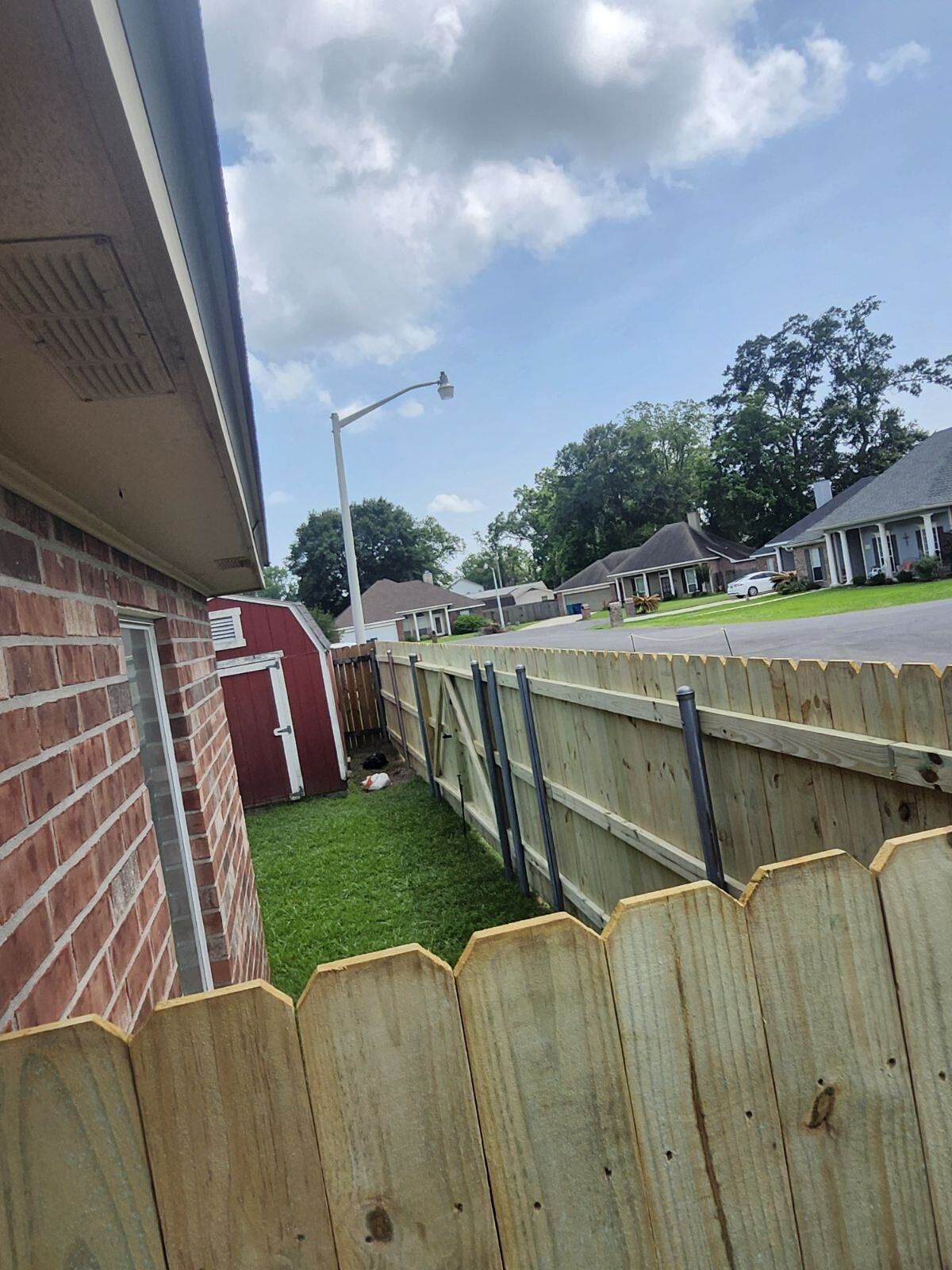 Narrow backyard between brick house and wooden fence, with grass, red shed, and cloudy sky.