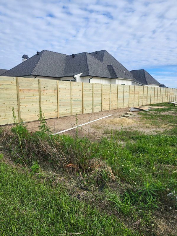 Long wooden fence beside a grassy lot, with a large gray-roofed house in the background.