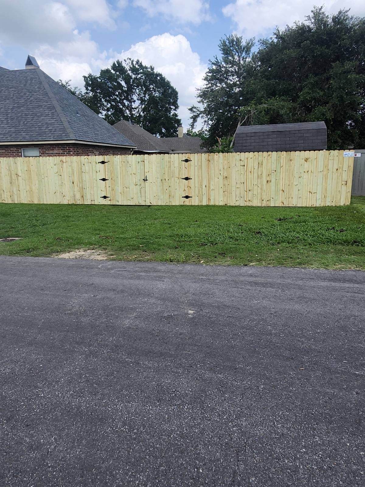 Wooden privacy fence beside a paved road, with grass and trees under a partly cloudy sky