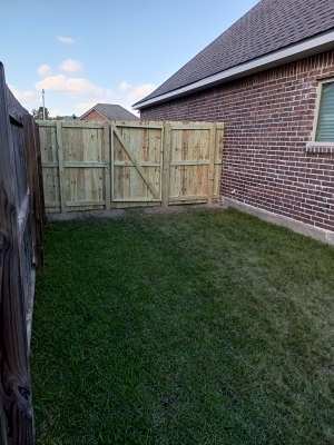 Backyard with green lawn, wooden gate, and brick house wall under a partly cloudy sky