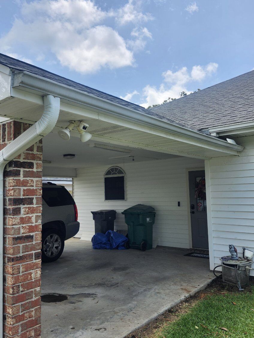 Residential carport with a parked SUV, trash bins, and blue bags under a white house roof.