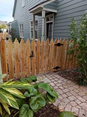 Wooden picket fence beside a gray house, with a paver walkway and leafy plants in the foreground.
