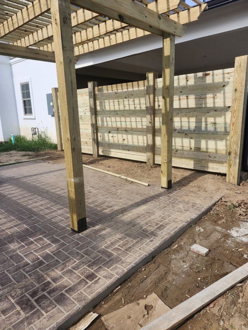 Wooden pergola over a paved walkway beside a fenced building courtyard.