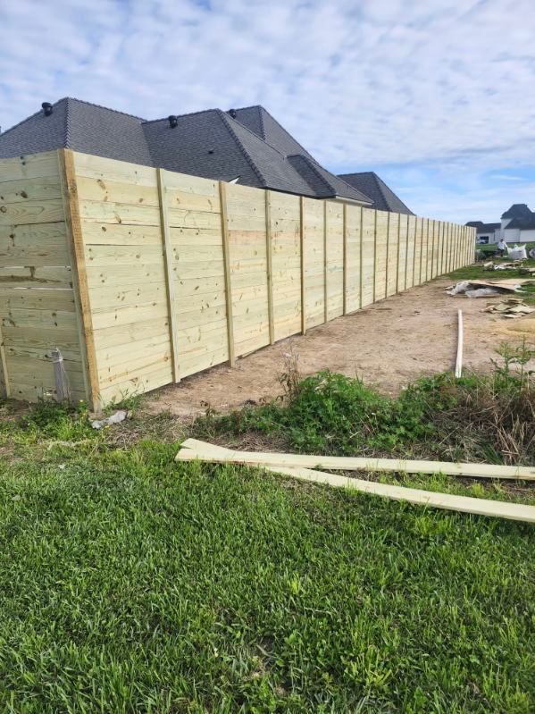 Long wooden fence panels line a yard beside houses, with grass and dirt in the foreground.