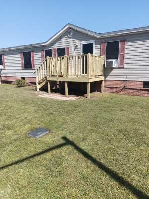 Mobile home with a small wooden front porch and red shutters on a grassy lawn under a clear sky