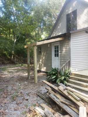 Exterior of a house with a white door, porch steps, stacked lumber, and trees in the yard