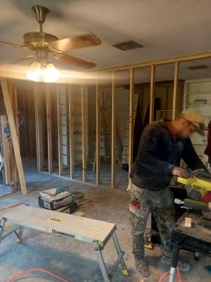 Person working with tools inside a room under renovation, with exposed wall studs and a ceiling fan.