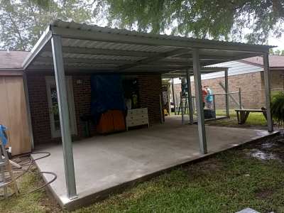 Covered patio with a concrete slab, metal posts, and a brick house beside a grassy yard.