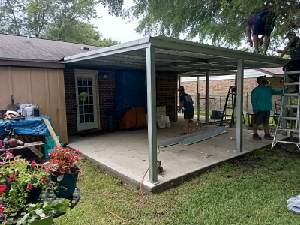 Covered patio beside a house, with a green post frame, blue wall, tools, and people working nearby
