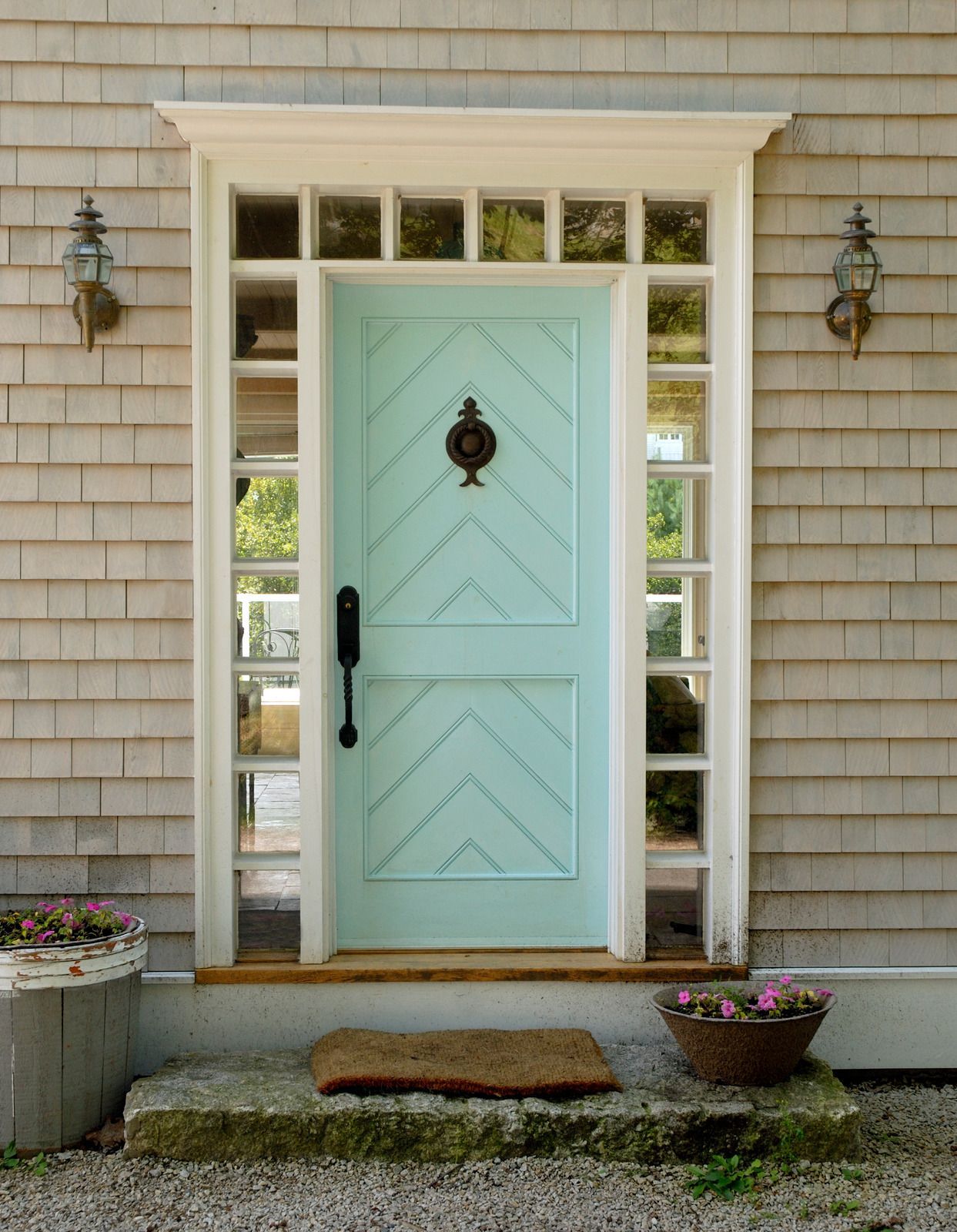 Light blue door with chevron design, flanked by sidelights. Two sconces, doormat, and flowerpots.