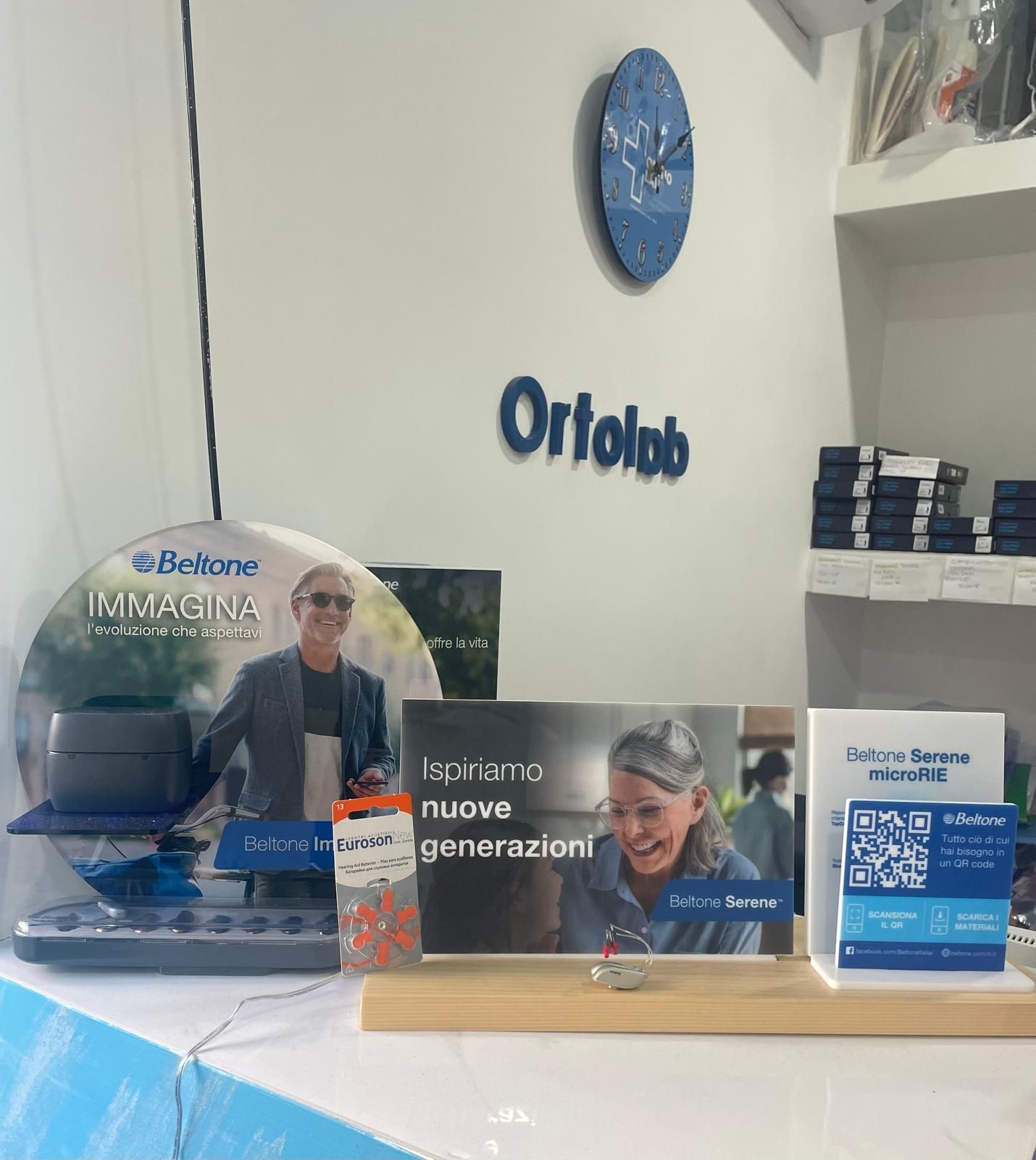 Espositore con prodotti per apparecchi acustici e materiale promozionale presso l'ufficio di Ortola. Include un orologio blu e il logo sulla parete bianca.