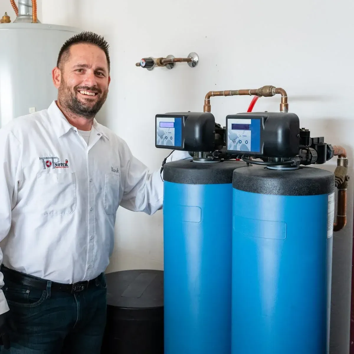 Technician standing beside two blue water treatment tanks with control units in a utility room Technician standing beside two blue water treatment tanks with control units in a utility room