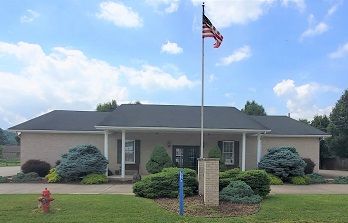 Single-story beige building with an American flag, trimmed shrubs, and a small lawn under a blue sky.