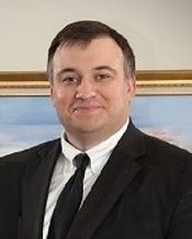 Man in a black suit and tie, smiling in a formal indoor portrait.