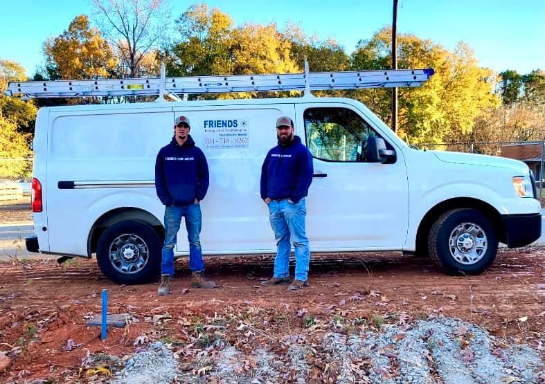 Two men in matching dark hoodies stand in front of a white work van with a ladder rack on an outdoor dirt lot.