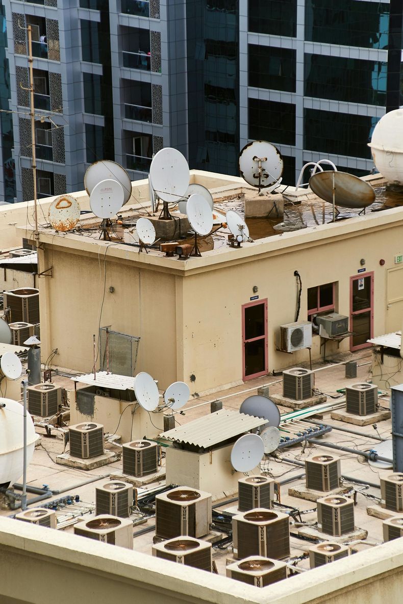 A rooftop filled with numerous air conditioning units and multiple satellite dishes against a background of tall buildings.