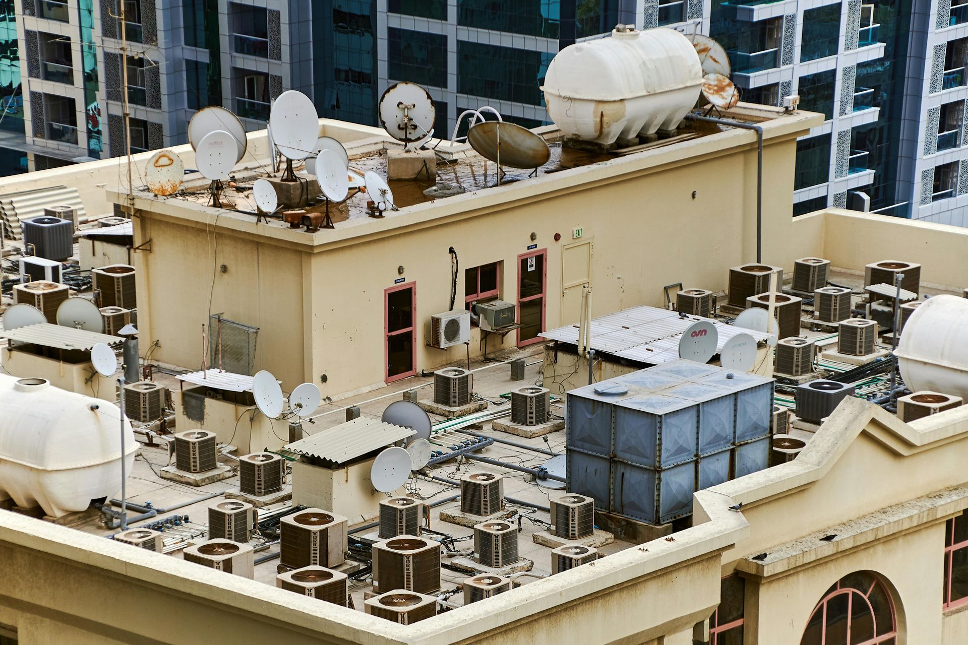A high-angle view of a beige building rooftop crowded with numerous air conditioning units, satellite dishes, and tanks.