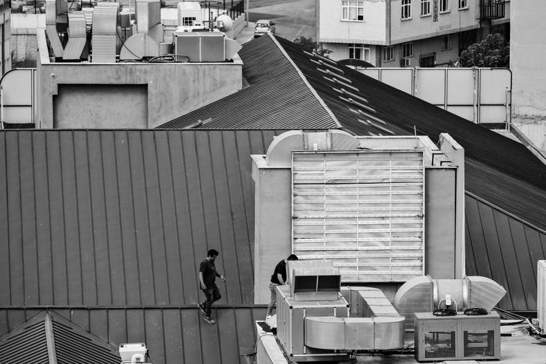 Two people walk on a sloped, metallic roof next to large industrial HVAC equipment.