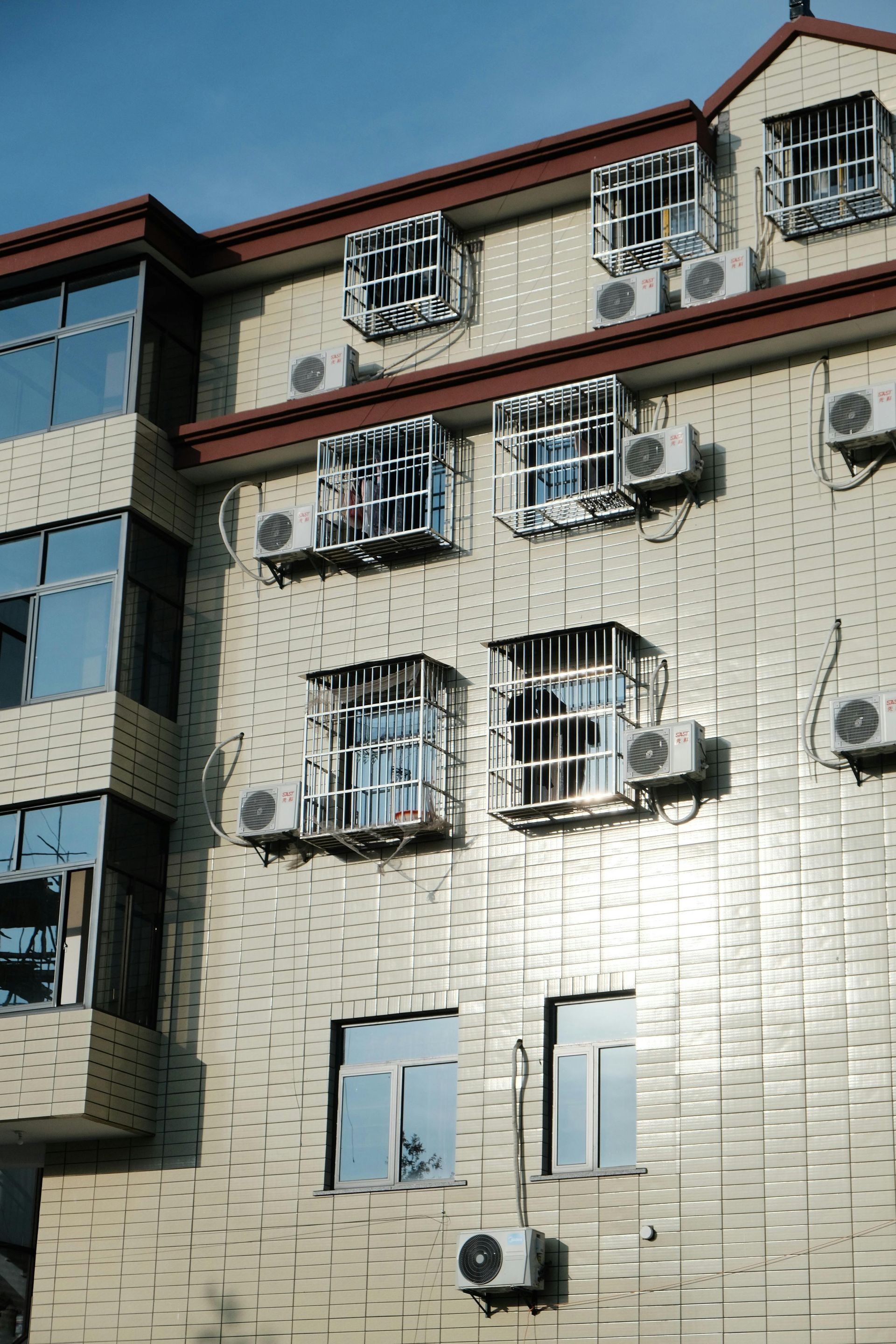 An exterior view of a building facade featuring light beige tiled walls, windows with white security grilles, and AC units.
