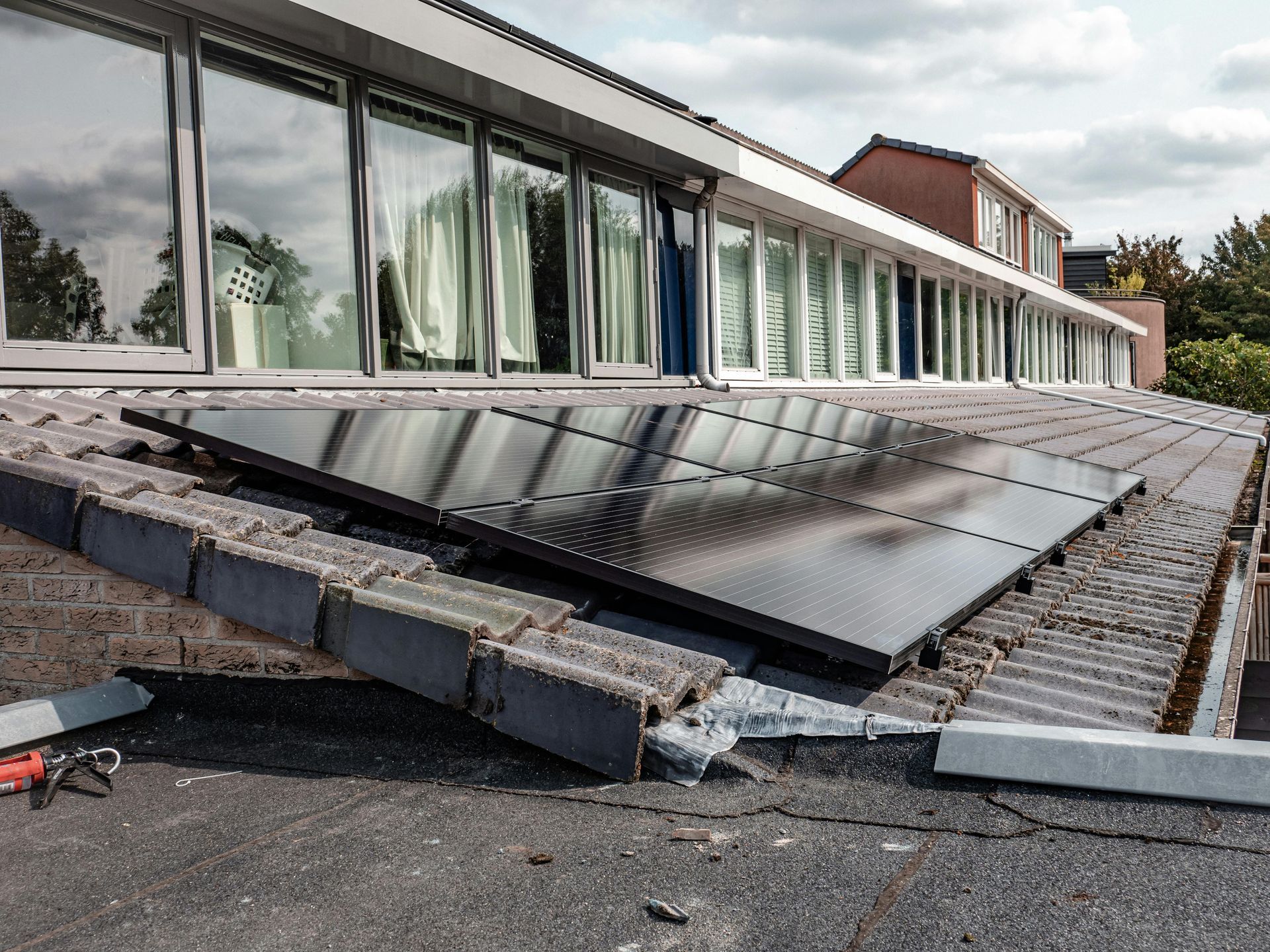 A black solar panel array installed on a grey, pitched tiled roof next to a modern building with large glass windows.