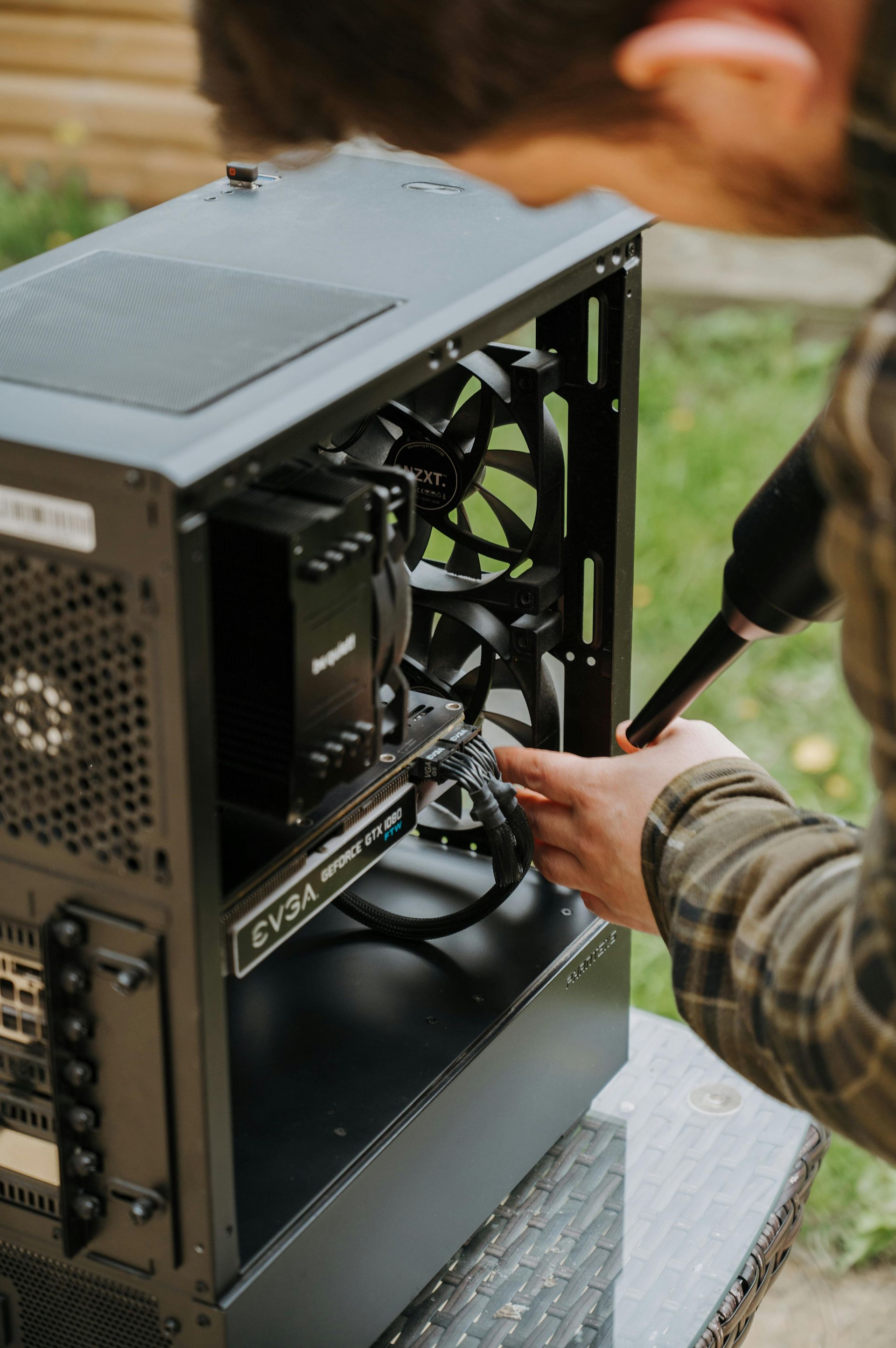 A person using an air duster to clean dust from the internal components of an open computer case outdoors.
