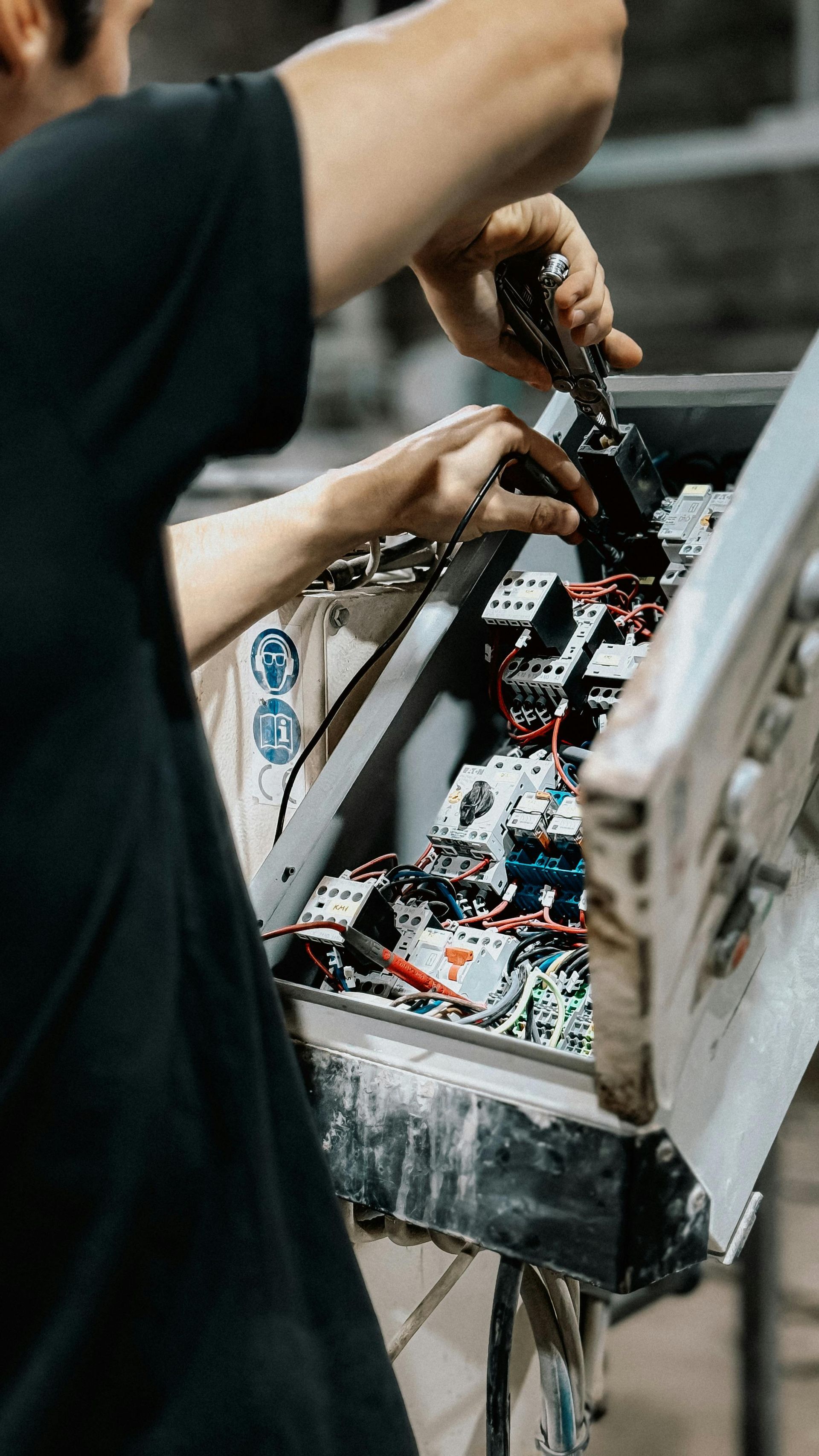 A person in a black t-shirt uses a screwdriver to work on the wiring inside an open electrical control panel.
