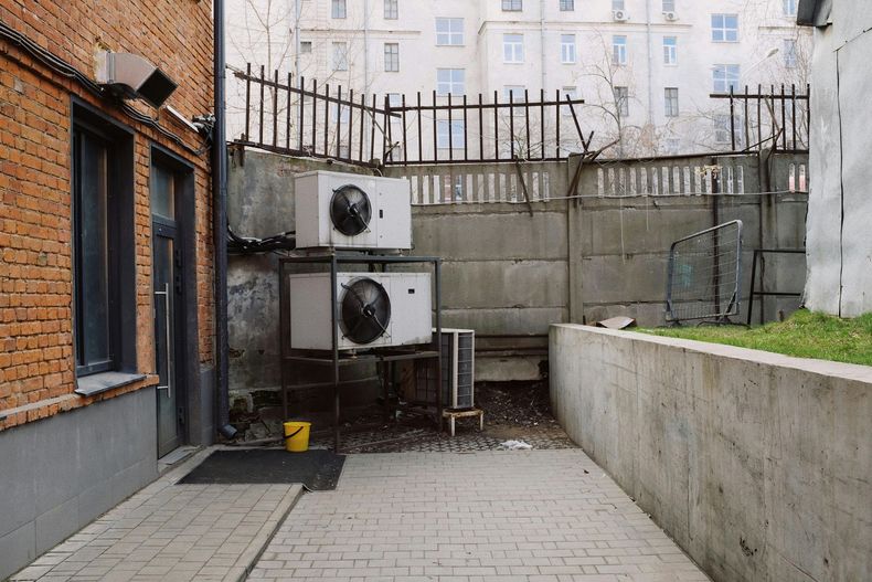 Two stacked air conditioner units sit on a metal frame against a concrete wall in a brick building's outdoor area.