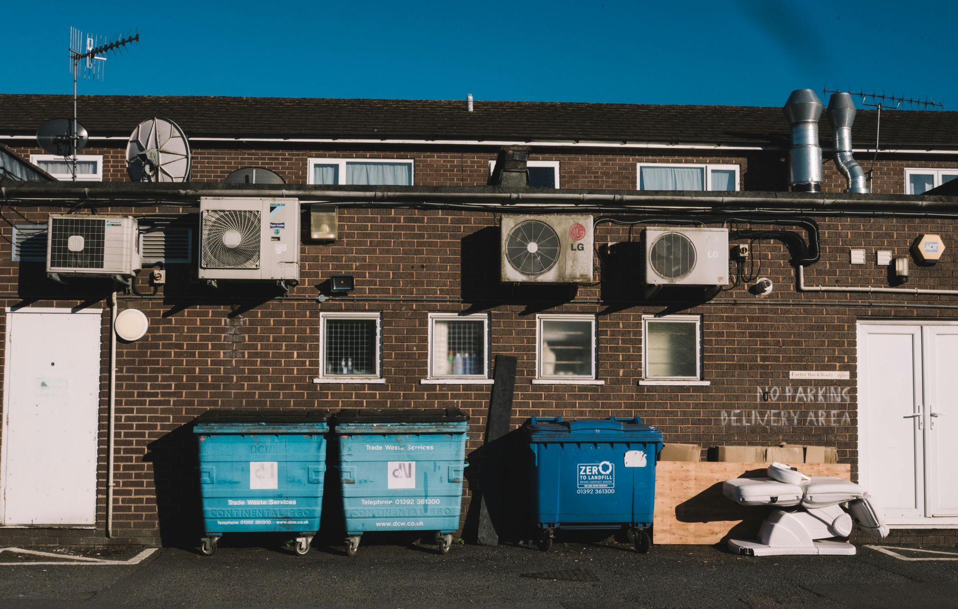 A brick building exterior with three blue dumpsters, outdoor HVAC units, and several windows under a clear blue sky.