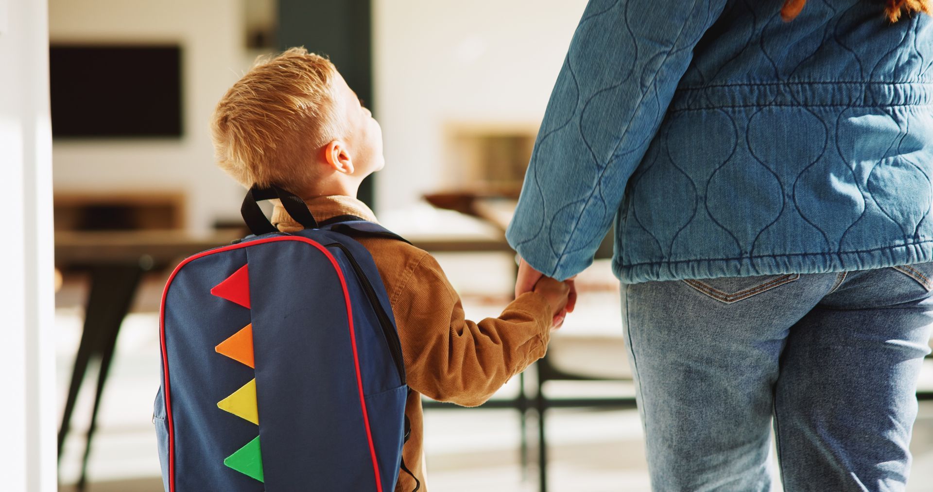 Person holding a child’s hand indoors, with the child wearing a colorful backpack.