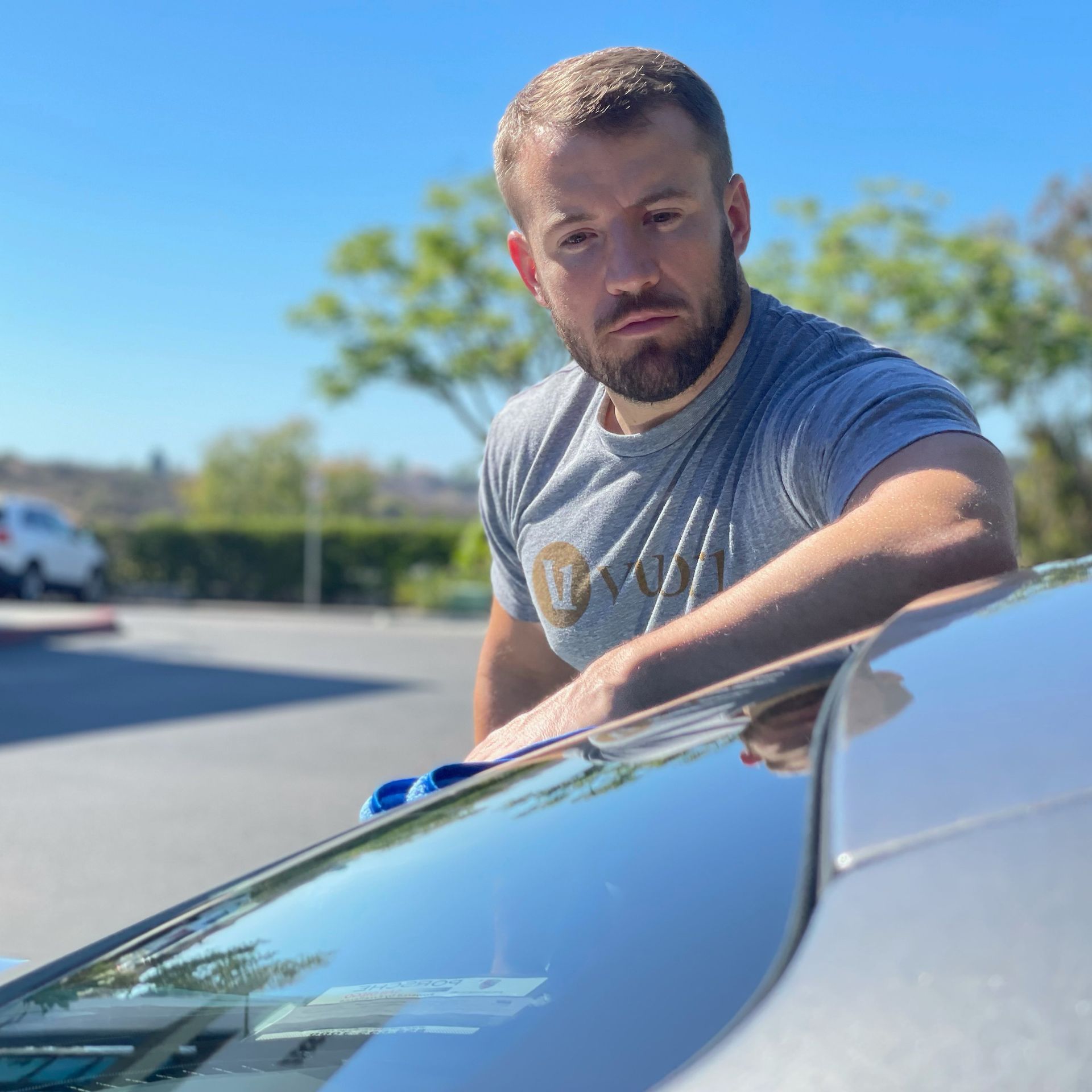 Man cleaning a car windshield on a sunny day; blue microfiber cloth, gray shirt, outdoor setting.