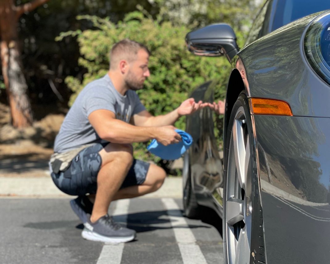 Man squats, wiping a gray sports car with a blue cloth in an outdoor setting.