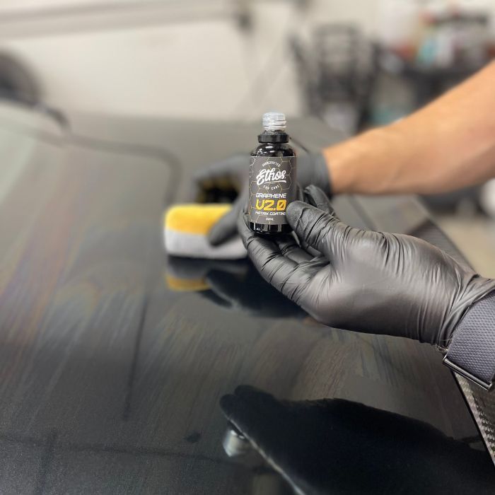 Person in black gloves applying car ceramic coating to a black car hood with a sponge.