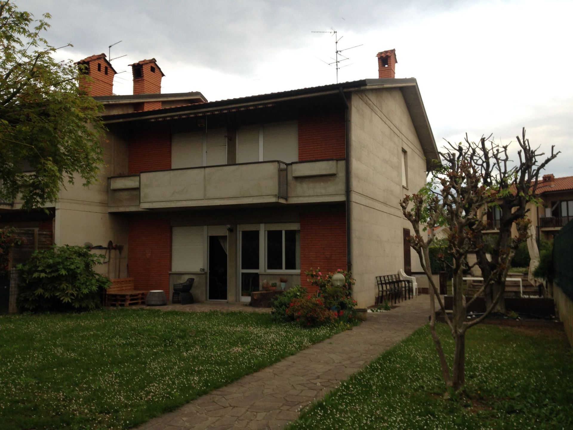 Casa beige a due piani con balcone, circondata da arbusti e una recinzione metallica, in una strada tranquilla.