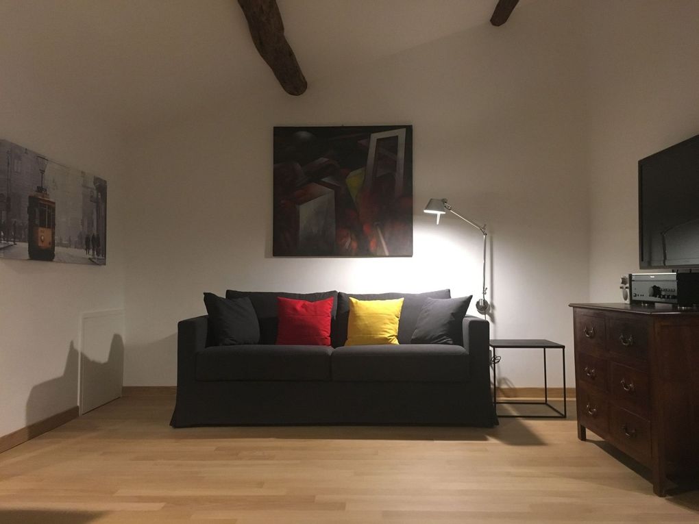 Soggiorno minimalista con divano scuro, cuscini colorati, lampada da terra, quadri e mobiletto in legno.