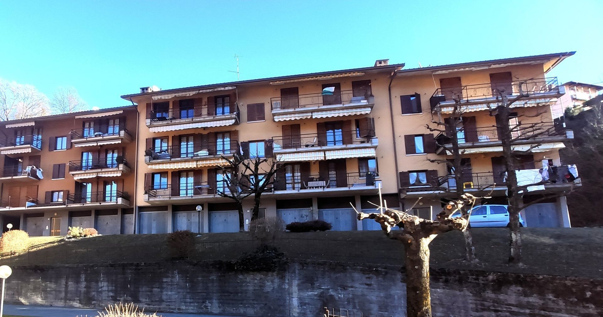 Una fila di palazzi marroni con balconi e un'auto parcheggiata dietro un muro di pietra in una giornata di sole.