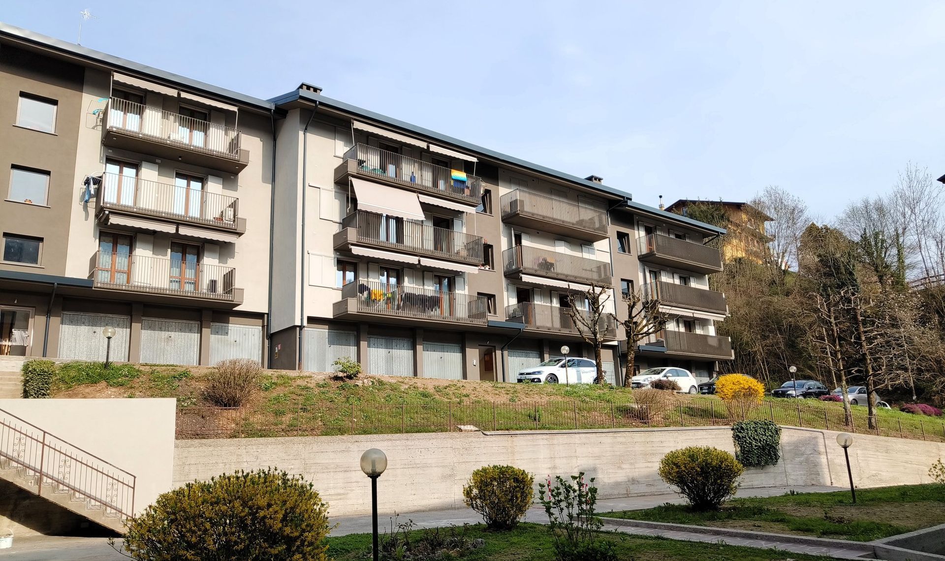 Condominio con balconi, vialetto alberato e arbusti in una giornata di sole.