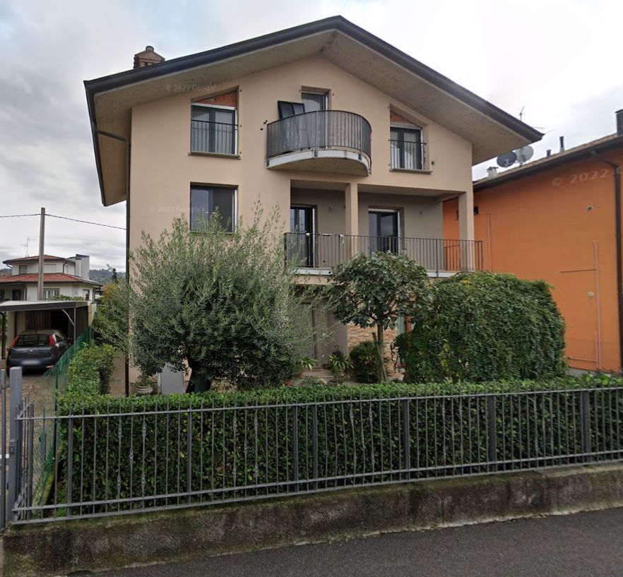 Casa beige a due piani con balcone, circondata da arbusti e una recinzione metallica, in una strada tranquilla.